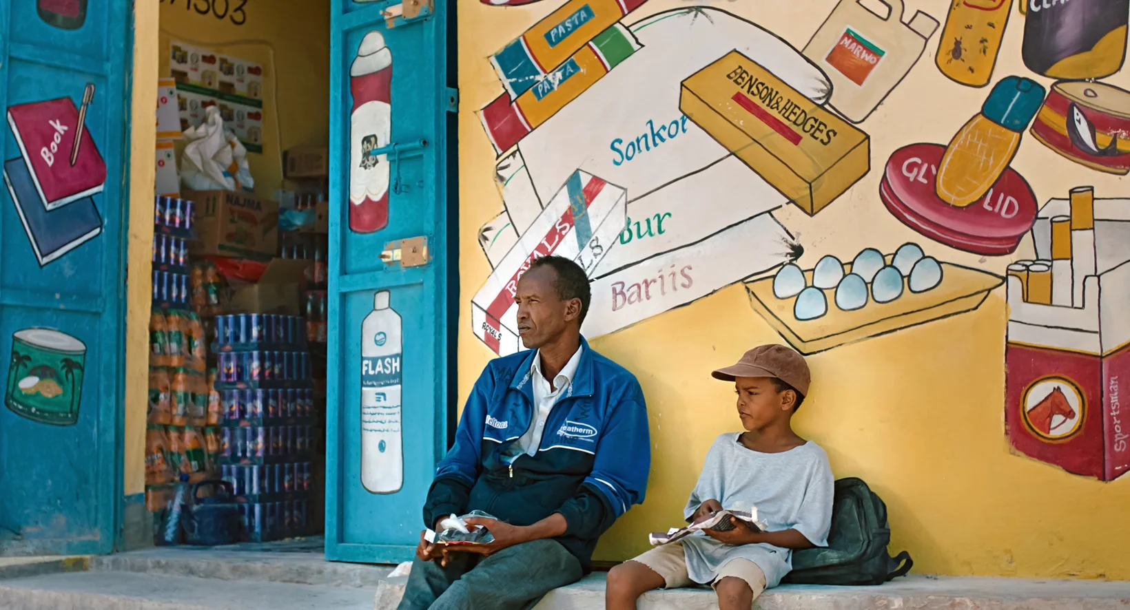 Vater und Sohn sitzen vor einer bunt bemalten Wand: Filmstill aus «The Village Next to Paradise»